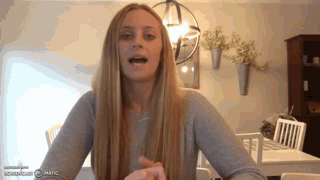 a woman with long blonde hair is sitting at a table in a living room with a chandelier in the background .