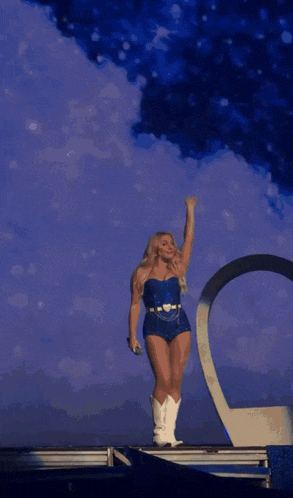 a woman in a blue bodysuit and white cowboy boots stands on stage