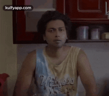 a man wearing a tank top with the word wellness on it is sitting in a kitchen .