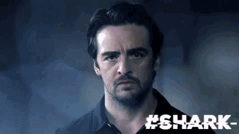 a man with a beard and a black shirt is looking at the camera with the words #shark behind him .