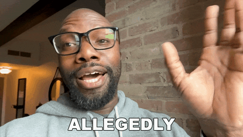 a man with glasses and a beard says allegedly in front of a brick wall