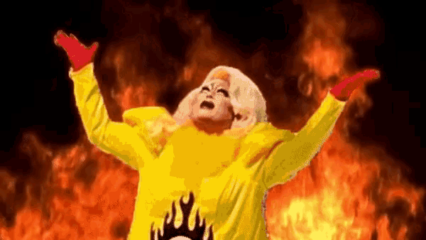 a drag queen in a yellow jacket is standing in front of a fire .