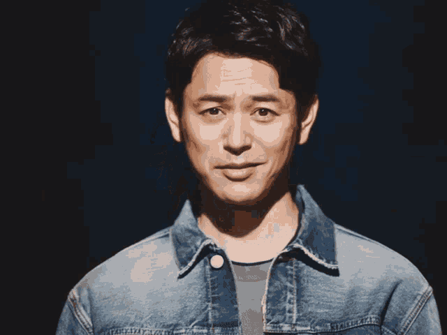 a man wearing a denim jacket and a grey shirt looks at the camera
