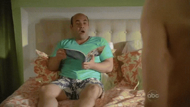 a man in a green shirt is laying on a bed reading a magazine ..