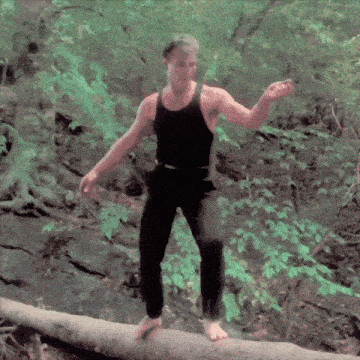 a man in a black tank top stands on a log in the woods
