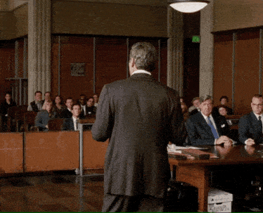 a man in a suit and tie stands in front of a group of people in a courtroom with an exit sign in the background