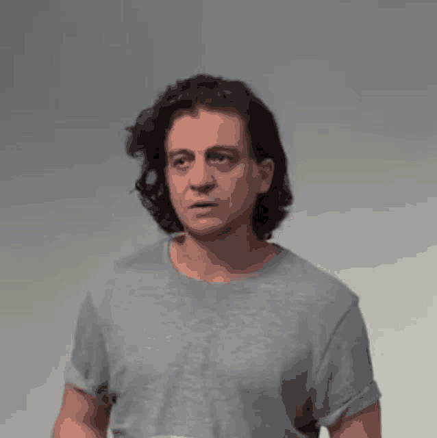 a man with long hair is wearing a gray t-shirt and looking at the camera .