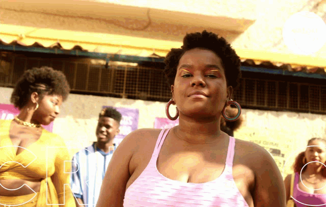 a woman in a pink tank top with hoop earrings stands in front of a group of people