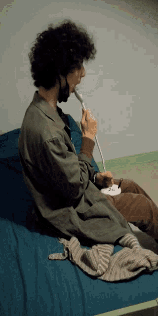 a man with curly hair is sitting on a bed with the number 26 written on a piece of paper