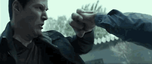 a man in a black jacket is giving another man a fist bump in front of a window .