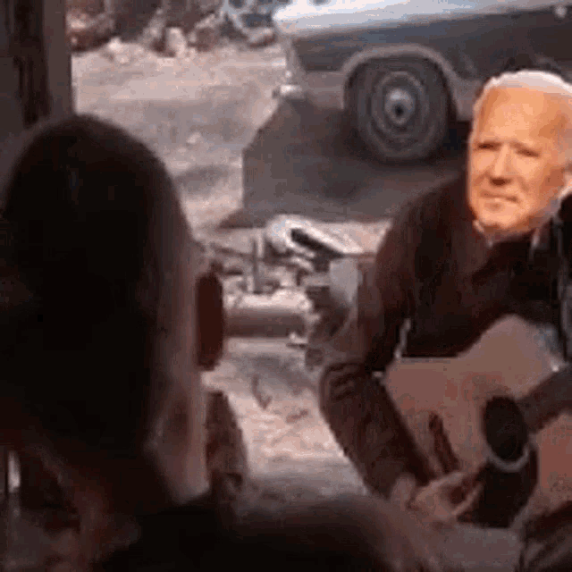 a man is playing a guitar in front of a car while another man looks on .