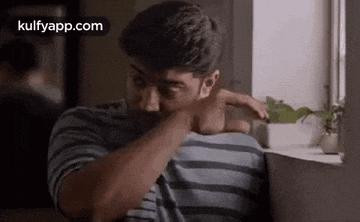 a man in a striped shirt is sitting in front of a window and rubbing his face .