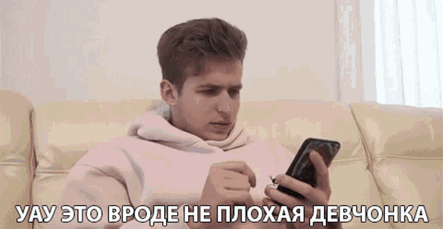 a man in a pink hoodie is sitting on a couch looking at his cell phone .