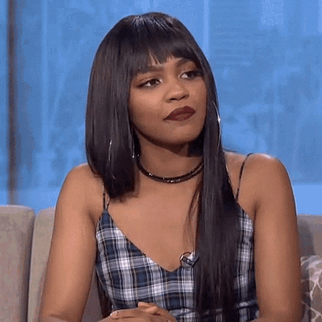 a woman is sitting on a couch wearing a plaid top and a choker .