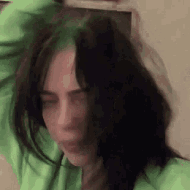 billie eilish is wearing a green hoodie and holding her hair .