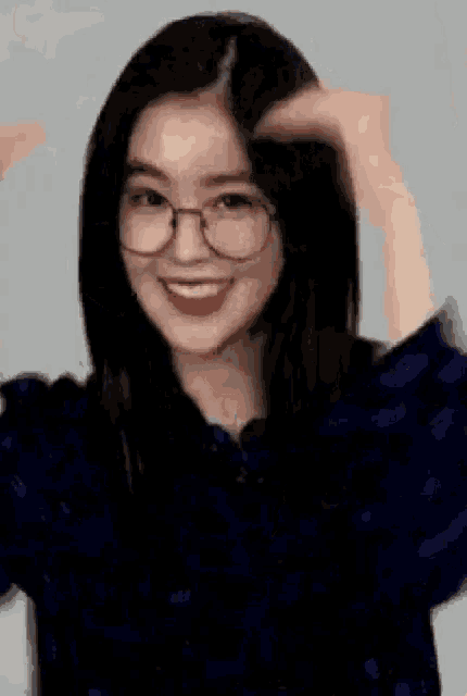 a young woman wearing glasses and a blue shirt is smiling and making a heart shape with her hands .
