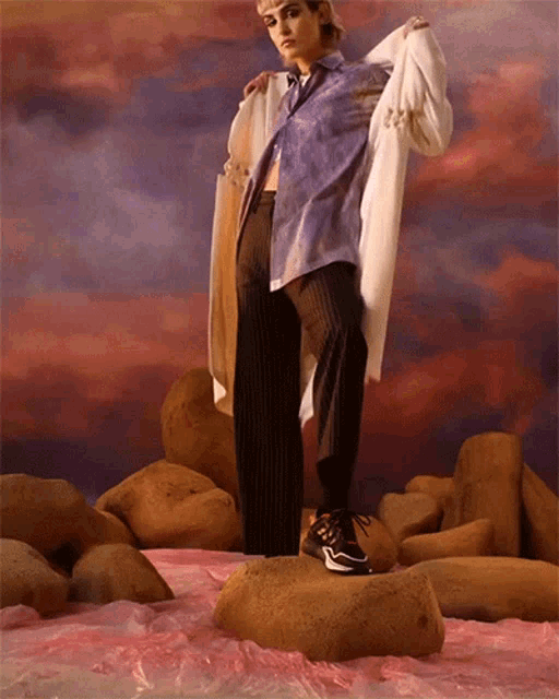 a woman in a purple shirt and black pants is standing on rocks