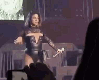 a woman is singing into a microphone on a stage while a person takes a picture with a cell phone .
