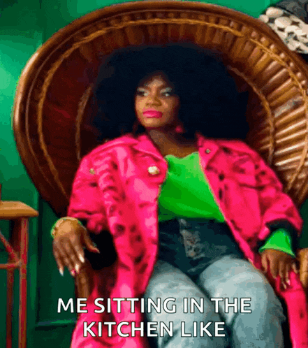 a woman in a pink jacket and green shirt is sitting in a wicker chair with the caption me sitting in the kitchen like