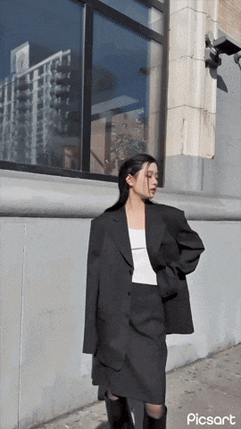 a woman wearing a black jacket and a white tank top is standing in front of a building with picsart written on the bottom