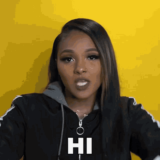 a woman wearing a black jacket says hi on a yellow background