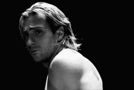 a man with long hair is wearing a white tank top and looking at the camera .