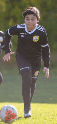 a young boy wearing a black adidas jersey and benjamin shorts runs towards a soccer ball