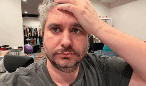 a man with a beard is holding his hand to his forehead and looking at the camera .