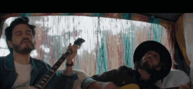 two men are playing guitars in a van with a colorful curtain behind them
