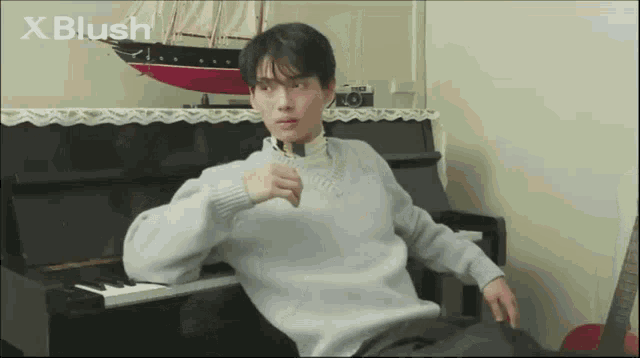 a man in a sweater sits in front of a piano with x blush written above him