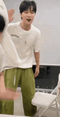 a man in a white t-shirt and green pants is standing next to a folding chair .