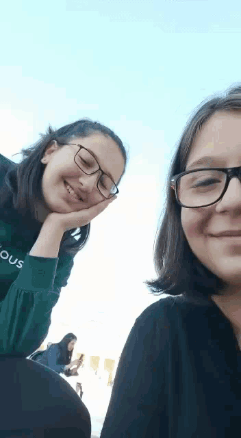 two girls wearing glasses are posing for a picture together . one girl is wearing a green sweatshirt that says famous .