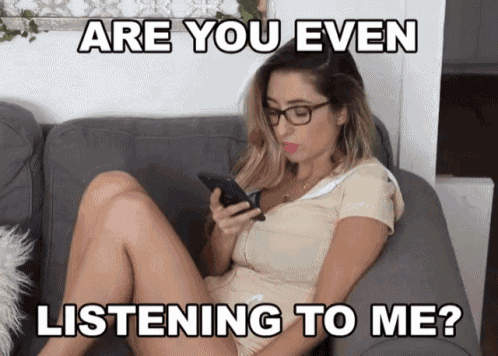 a woman sits on a couch looking at her phone with the caption " are you even listening to me " above her