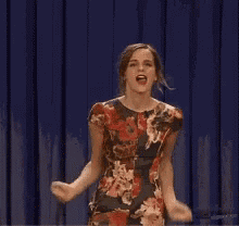 a woman in a floral dress is standing in front of a blue curtain on a stage .