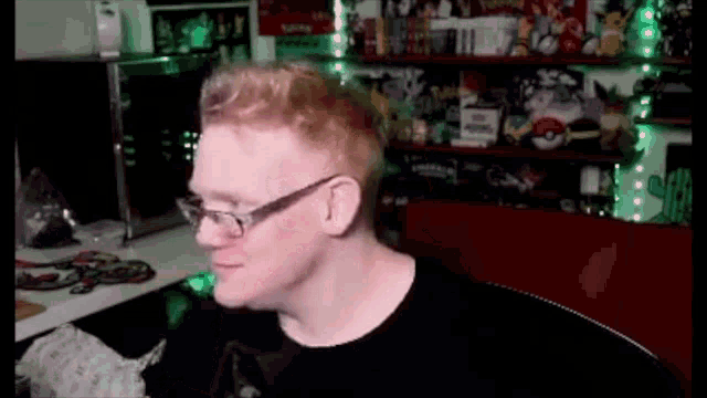 a man with red hair and glasses is sitting at a desk in front of a shelf filled with toys .