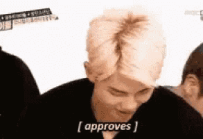 a man with blonde hair is making a funny face and saying `` approves '' while sitting in front of a white background .