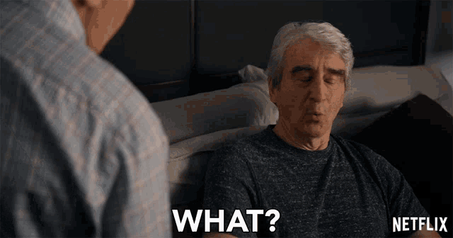 an older man is sitting on a couch talking to another man and the man is asking " what "