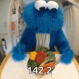 cookie monster from sesame street is sitting at a table with a plate of vegetables in front of him .