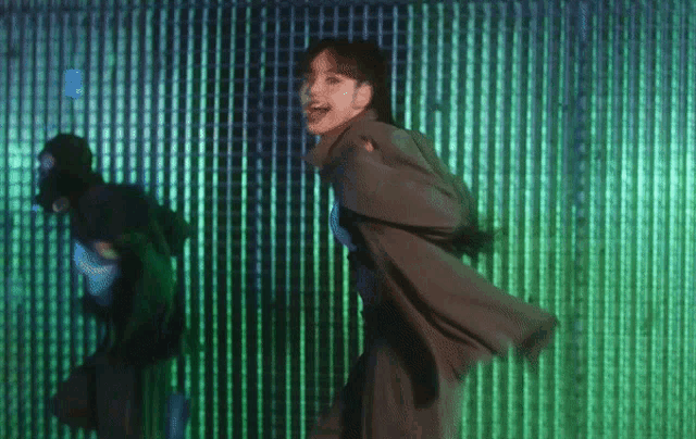 a woman in a trench coat is dancing in front of a green wall