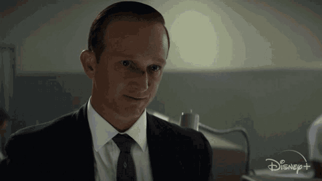 a man in a suit and tie is sitting in front of a disney+ logo