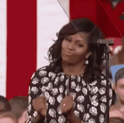 michelle obama is standing in front of a microphone in front of a crowd and clapping her hands .