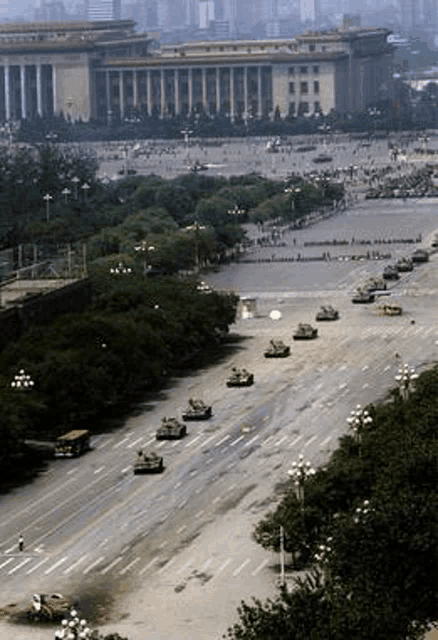 a row of tanks are driving down a city street in front of a building .