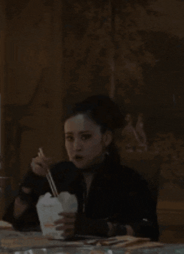 a woman is sitting at a table eating noodles with chopsticks from a takeout box .