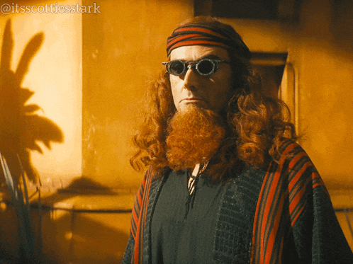 a man with long red hair and a beard wears sunglasses and a headband