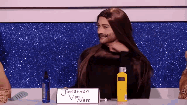 a man with long hair is sitting at a table with a sign that says jonathan van ness on it
