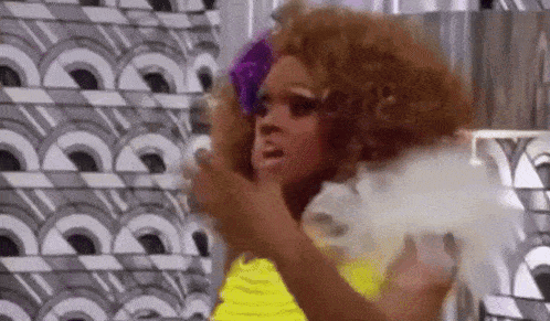 a drag queen with a purple flower in her hair and a yellow dress is standing in front of a patterned wall .