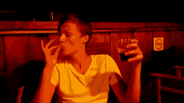 a man smoking a cigarette while holding a glass of beer in front of a no smoking sign