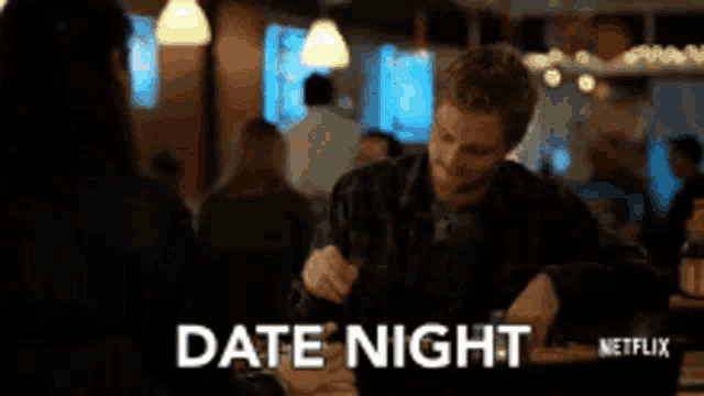 a man is sitting at a table with a glass of wine and the word date night is on the screen behind him .