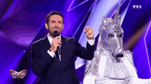 a man in a suit holds a microphone next to a masked unicorn