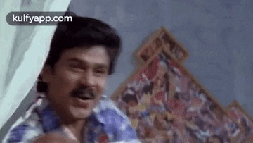 a man with a mustache is sitting on a bed with a painting on the wall behind him .